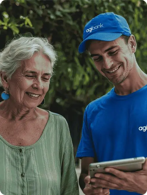 Consultor do Agibank, vestindo camiseta e boné azuis com o logo da empresa, mostra informações em um tablet para uma mulher idosa sorridente, em ambiente externo durante o dia.