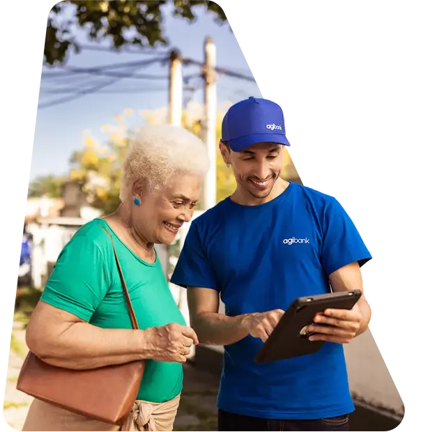 Consultor do Agibank, vestindo camiseta e boné azuis com o logo da empresa, mostra informações em um tablet para uma mulher idosa sorridente, em ambiente externo durante o dia.