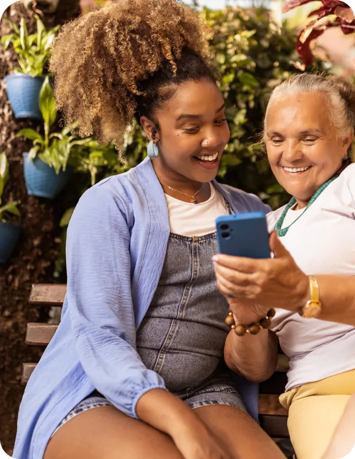 Duas mulheres sorrindo enquanto olham para um celular juntas em um ambiente com plantas.